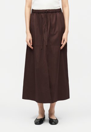 Žena nosi tmavohnedú midi sukňu so sťahovacou šnúrkou v páse a čierne balerínky, stojaca na svetlom pozadí.