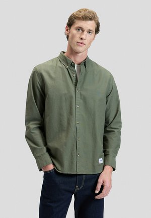 Młody mężczyzna o jasnobrązowych włosach, ubrany w oliwkowo-zieloną koszulę na guziki na biały podkoszulek i ciemnoniebieskie dżinsy na jednolitym tle.