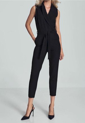 Femme portant une combinaison noire sans manches avec ceinture, jambes resserrées et escarpins noirs pointus, debout devant un fond uni.
