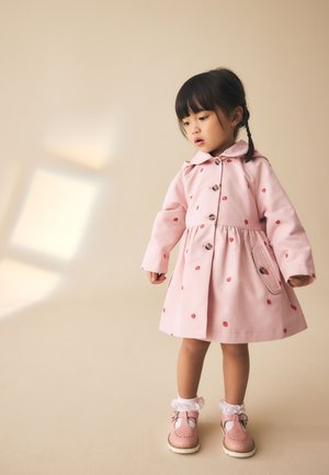 Junges Mädchen mit geflochtenen Haaren, das einen rosa Mantel mit roten Mustern, weiße Spitzenstrümpfe und rosa Schuhe trägt, steht vor einem beigefarbenen Hintergrund.