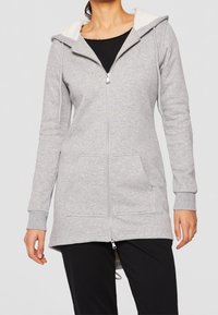 Donna che indossa una felpa grigia con cerniera e cappuccio, con tasche frontali, sopra una maglia nera e pantaloni neri, in piedi con le braccia rilassate.