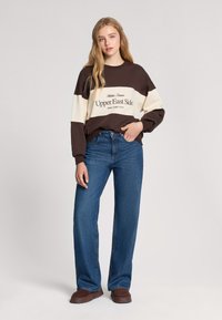 Felpa a maniche lunghe marrone e crema con il testo "Upper East Side", abbinata a jeans a gamba larga blu e scarpe slip-on marroni.