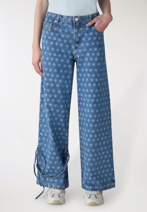 Jeans di denim a gamba larga blu con leggero motivo a pois, cinturini laterali e tasche frontali, indossati con sneakers bianche e top infilato dentro.