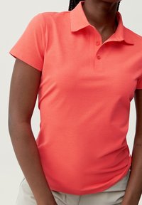 Korallfärgad pikétröja med korta ärmar och en mjuk textur, med krage och tre knappar vid knappslån.
