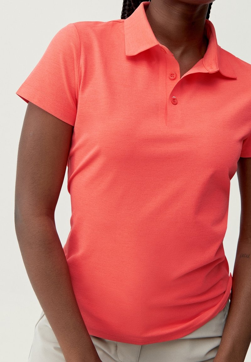 Korallfärgad pikétröja med korta ärmar och en mjuk textur, med krage och tre knappar vid knappslån.