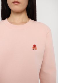 Femme portant un sweat-shirt rose pâle avec un petit motif brodé de gâteau à la fraise sur la poitrine, sur un fond gris uni.