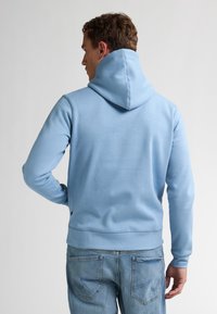 Sudadera azul claro hecha de una tela suave, con una capucha grande, mangas largas y un cinturón ajustado, combinada con jeans.