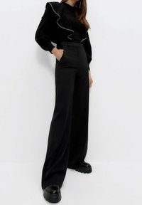 Černý top s volány spárovaný s vysokými černými kalhotami s širokými nohavicemi. Modelka má na sobě masivní černé platformové boty.