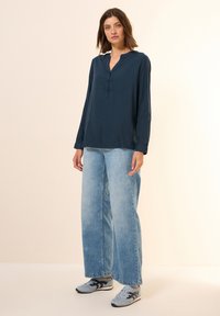Marineblauwe blouse met lange mouwen en een gedeeltelijke knoopsluiting, gecombineerd met lichtblauwe wijde jeans en grijze sneakers met zwarte accenten.