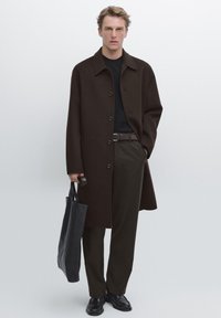 Cappotto in lana marrone con bottoni, dolcevita nera, pantaloni verde scuro e scarpe in pelle nere; tiene una borsa a tracolla nera.