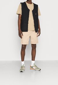 Muž v béžovém tričku a šortkách, který nosí černou bezrukávovou pracovní vestu s kapsami, bílé ponožky a béžovo-zelené tenisky New Balance.