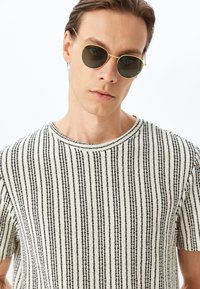 Camiseta de punto a rayas blancas y negras con cuello redondo. La modelo lleva gafas de sol redondas doradas. Fondo blanco.
