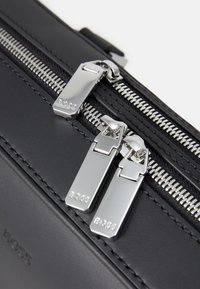 Sac en cuir noir avec deux zips argentés arborant le nom de la marque. Fini texturé, design épuré et coutures visibles le long des bords.