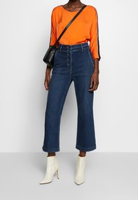 Oranges Langarmoberteil mit schwarzen Akzenten, kombiniert mit hochtaillierten dunkelblauen Denim-Culottes und weißen Ankle-Boots, dazu eine schwarze Umhängetasche.