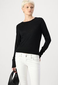 Svart langermet genser med teksturdesign prydet med små detaljer. Kombinert med hvite jeans og en svart håndveske.