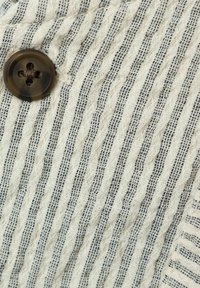 Tessuto a righe con una finitura texture in crema e blu scuro. Presenta un bottone marrone con quattro fori.