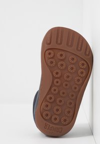 Suola di una scarpa per bambini, in gomma marrone con modelli di presa circolari. La vista laterale mostra una parte superiore blu navy con una finitura testurizzata e dettagli azzurri.