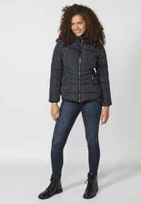 Padded navy blue jacket with high collar, vertical quilting, and gold zipper. Worn over dark denim jeans and black lace-up boots.
