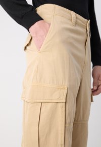 Béžové cargo nohavice s voľným strihom, ktoré majú bočné vrecká, šité detaily a zips na prednej strane. Materiál sa zdá byť ľahký a odolný.