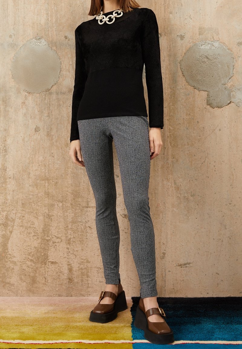 Femme portant un haut noir à manches longues, un collier chaîne blanc épais, un pantalon slim gris à motifs et des chaussures compensées marron avec boucles.