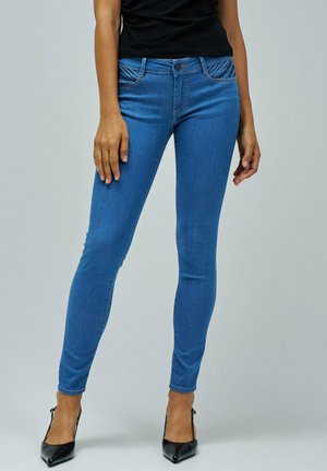 Jean bleu skinny avec taille mi-haute, présentant des coutures détaillées et des poches, associés à des chaussures à talons noirs.