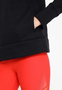 Sweatshirt noir avec poignets côtelés, doté d'une poche latérale. Associé à des leggings rouges arborant un motif en maille géométrique.