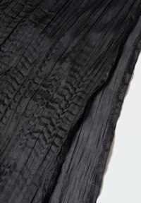 Tissu noir froissé avec des plis verticaux visibles, texture douce et panneau légèrement transparent le long d'un bord sur un fond blanc.