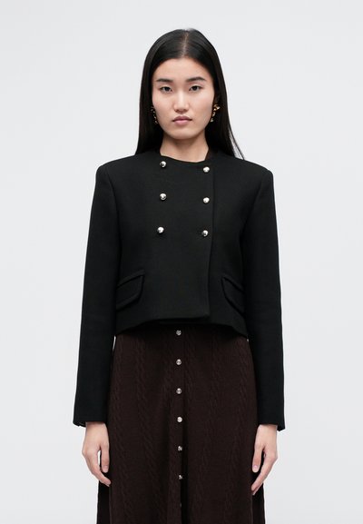 Schwarze, kurze Jacke aus strukturiertem Stoff mit silbernen Knopf-Details und zwei Fronttaschen. Getragen über einem dunkelbraunen Strickrock.