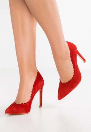 Rode suède hoge hakken met gegolfde randen, spitse neuzen en slanke stiletto-hakken. Gladde textuur en levendige kleur.