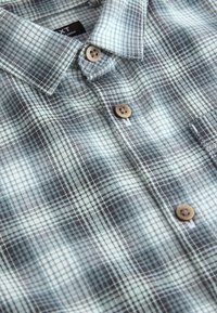 Chemise en flanelle aux nuances de bleu et gris avec un motif à carreaux, dotée d'un col et de boutons marron. Texture douce avec une coupe décontractée.