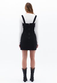 Schwarzes Denim-Kleid mit verstellbaren Trägern, Reißverschluss auf der Rückseite und tailliertem Schnitt. Getragen über einem weißen Langarmhemd. Schwarze Stiefel.