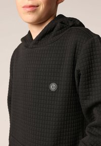 Zwarte hoodie met een doorgestikte textuur, een ronde logopatch op de borst en een capuchon met trekkoord, gemaakt van zachte stof.
