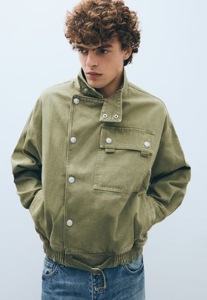 Jeune homme aux cheveux bouclés portant une veste boutonnée vert olive avec une poche poitrine et un jean bleu, mains dans les poches, regardant sur le côté.