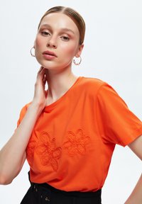 Camiseta de color naranja brillante con tres diseños de flores bordados. Mangas cortas, tejido suave, combinada con un cinturón negro de cintura alta.