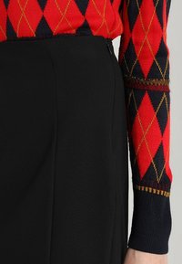 Gros plan sur une personne portant un pull argyle rouge et marine rentré dans un pantalon noir taille haute avec une fermeture éclair sur le côté.