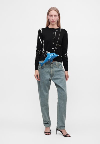 Femme portant un cardigan noir boutonné, un jean bleu clair ample, des talons noirs et un sac bandoulière bleu en forme de dauphin sur une chaîne.