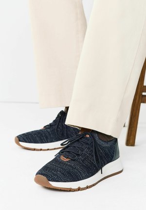 Person wearing navy knit sneakers with white soles and beige pants, seated on a wooden chair against a white background.
