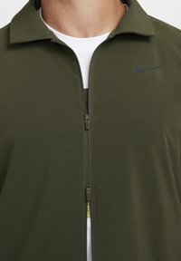 Alyvuogių žalios spalvos zip-up striukė su apykakle, pagaminta iš lygaus audinio. Savyje turi juodą Nike logotipą krūtinėje ir priekinį užtrauktuką.