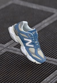 Blue and gray athletic sneaker with white sole and "N" logo, floating above a metal grid surface.
