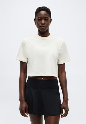 Νεαρό άτομο με κοντά σγουρά μαλλιά φοράει ένα λευκό κοντό T-shirt και μαύρη φούστα, στέκεται μπροστά σε απλό φωτεινό φόντο.