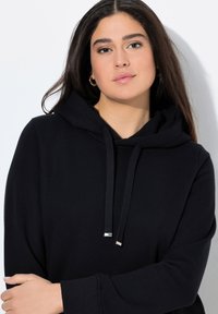 Sudadera negra con capucha de textura suave, que presenta un bolsillo canguro, detalles en metal plateado en los cordones y un corte relajado.