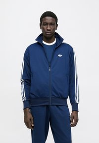 Μπλε ναυτικό τζάκετ με φερμουάρ και ψηλό γιακά, με λευκές τρεις ρίγες στα μανίκια και μικρό λευκό λογότυπο στο στήθος. Λείος υφιστάμενος.