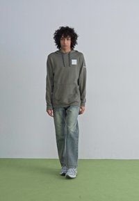 Felpa verde oliva con tasca frontale, cappuccio con cordini e etichetta; abbinata a jeans chiari di denim e scarpe grigie su un pavimento verde.