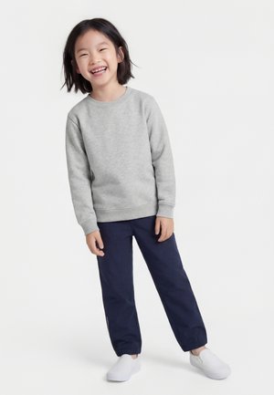 Copil zâmbitor cu păr scurt și închis la culoare, purtând un hanorac gri deschis, pantaloni bleumarin cu manșete elastice și pantofi albi slip-on, stând pe un fundal alb.