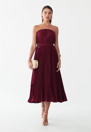 Vrouw draagt een strapless, geplooide bordeaux midi-jurk met een tailleriem, houdt een beige clutch vast en draagt gouden oorbellen en hakken.