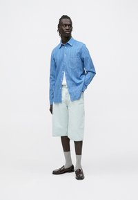 Mężczyzna o ciemnej karnacji ubrany w niebieską koszulę w paski na guziki, jasne szorty dżinsowe, szare skarpetki i ciemnobrązowe loafersy, stojący z jedną ręką w kieszeni.