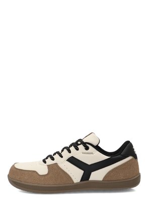 Zapatilla baja con ante beige y paneles de cuero crema, logo lateral negro, cordones negros y suela de goma marrón.