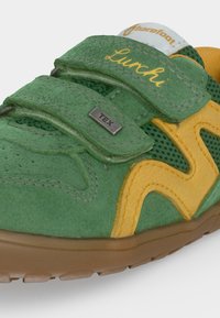 Chaussure pour enfant verte et jaune avec deux bandes velcro, étiquettes "Lunchi" et "Tex", et une semelle en caoutchouc marron.