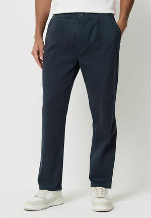 Navyblaue Hosen aus glattem Stoff, mit flachem Vorderteil, seitlichen Taschen und elastischem Bund, kombiniert mit weißen Sneakers.