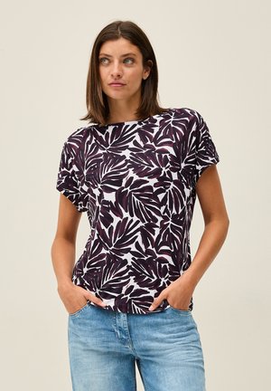 Top de mujer de manga corta en blanco con un estampado de hojas en púrpura oscuro. Cuello redondo, corte relajado y tela suave. Combinado con jeans azules claros.